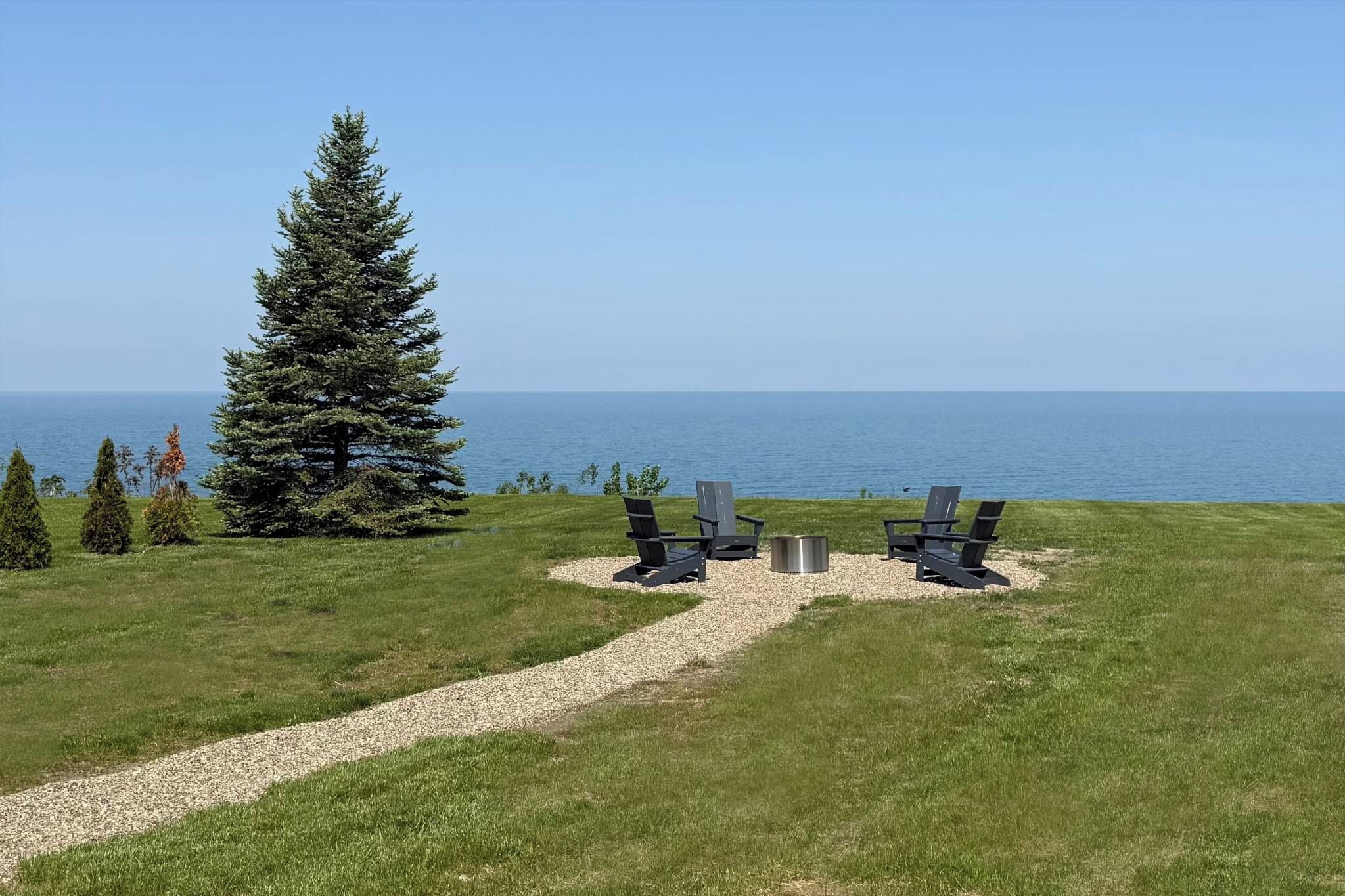 A grassy area with a gravel path leading to two chairs and a fire pit, overlooking a body of water.