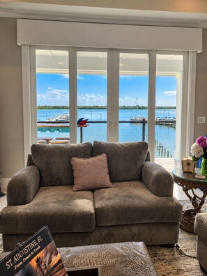 A living room with a large window overlooking a marina, a sofa, and a small table with a vase. - The Grand Mariner: Waterfront + Boat Slip + Elevator Luxury Estate (St. Augustine)