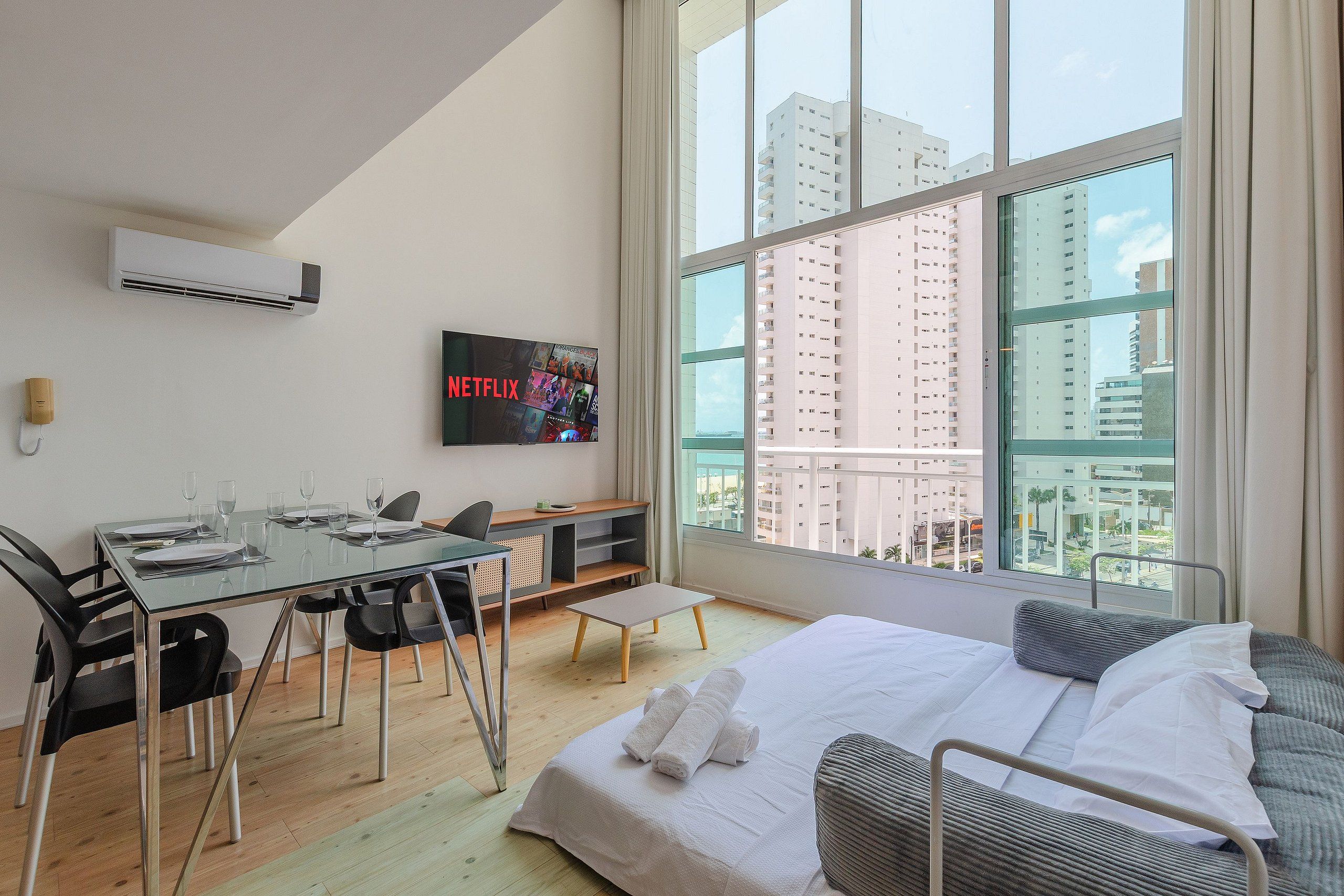 Habitación de hotel moderna con un ventanal grande, una cama, un escritorio con sillas, un televisor de pantalla plana y un equipo de aire acondicionado.