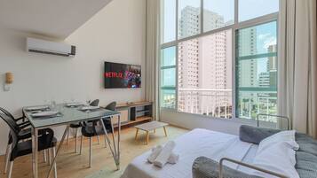 Habitación de hotel moderna con un ventanal grande, una cama, un escritorio con sillas, un televisor de pantalla plana y un equipo de aire acondicionado.