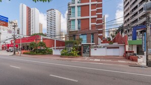 A street view with a building featuring a red facade, a gated entrance, and a small garden area. - Loft Praia #502 Loft With Sea View by Carpediem (Fortaleza)