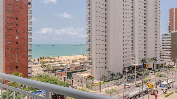A high-rise building with multiple balconies overlooking a beach and a busy street. - Loft Praia #502 Loft With Sea View by Carpediem (Fortaleza)