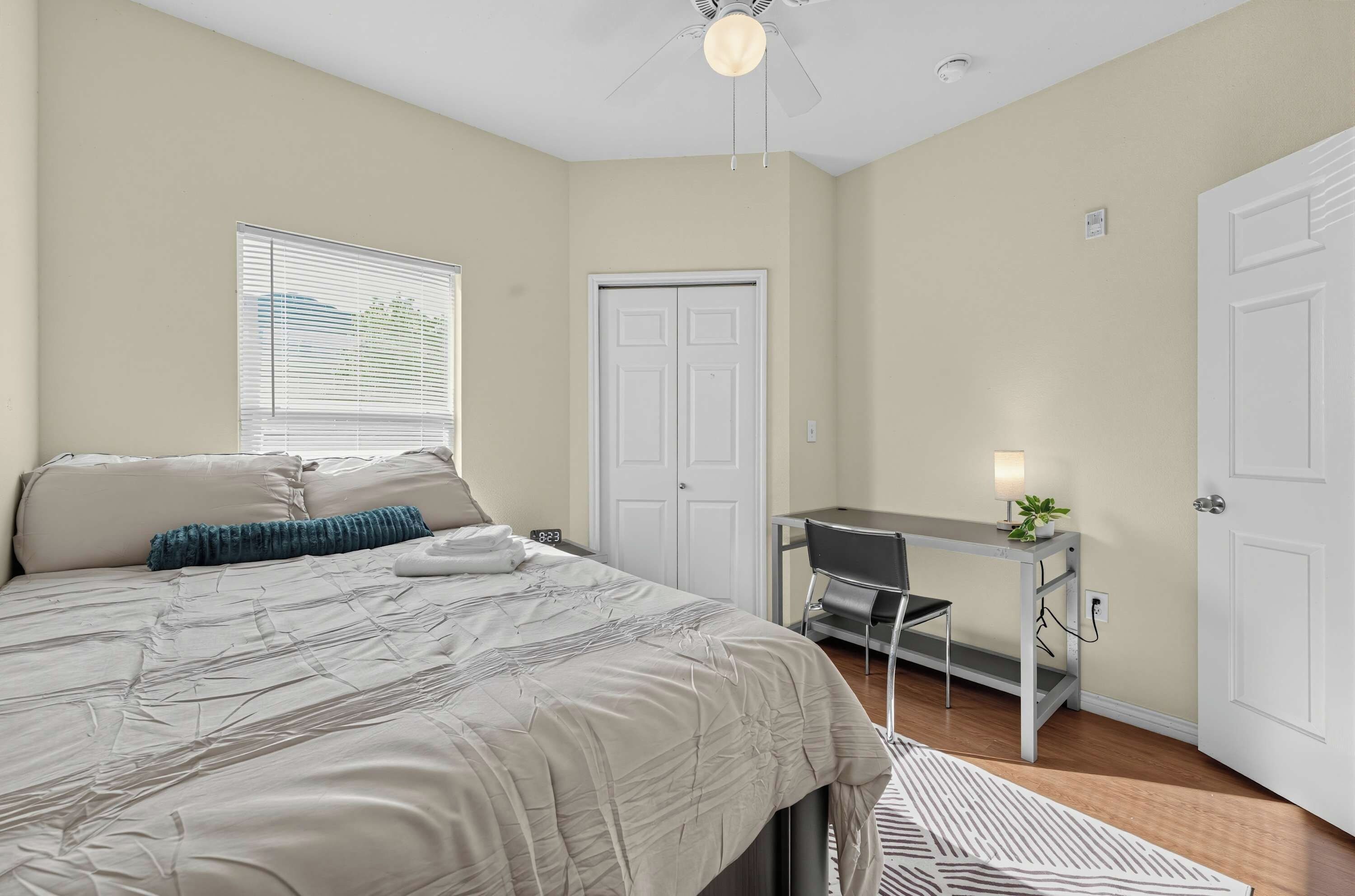 A bedroom with a bed, a desk, a chair, a window with blinds, and a door.
