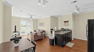 A modern kitchen with a central island, a dining area with a table and chairs, and a living room with a sofa and a ceiling fan.