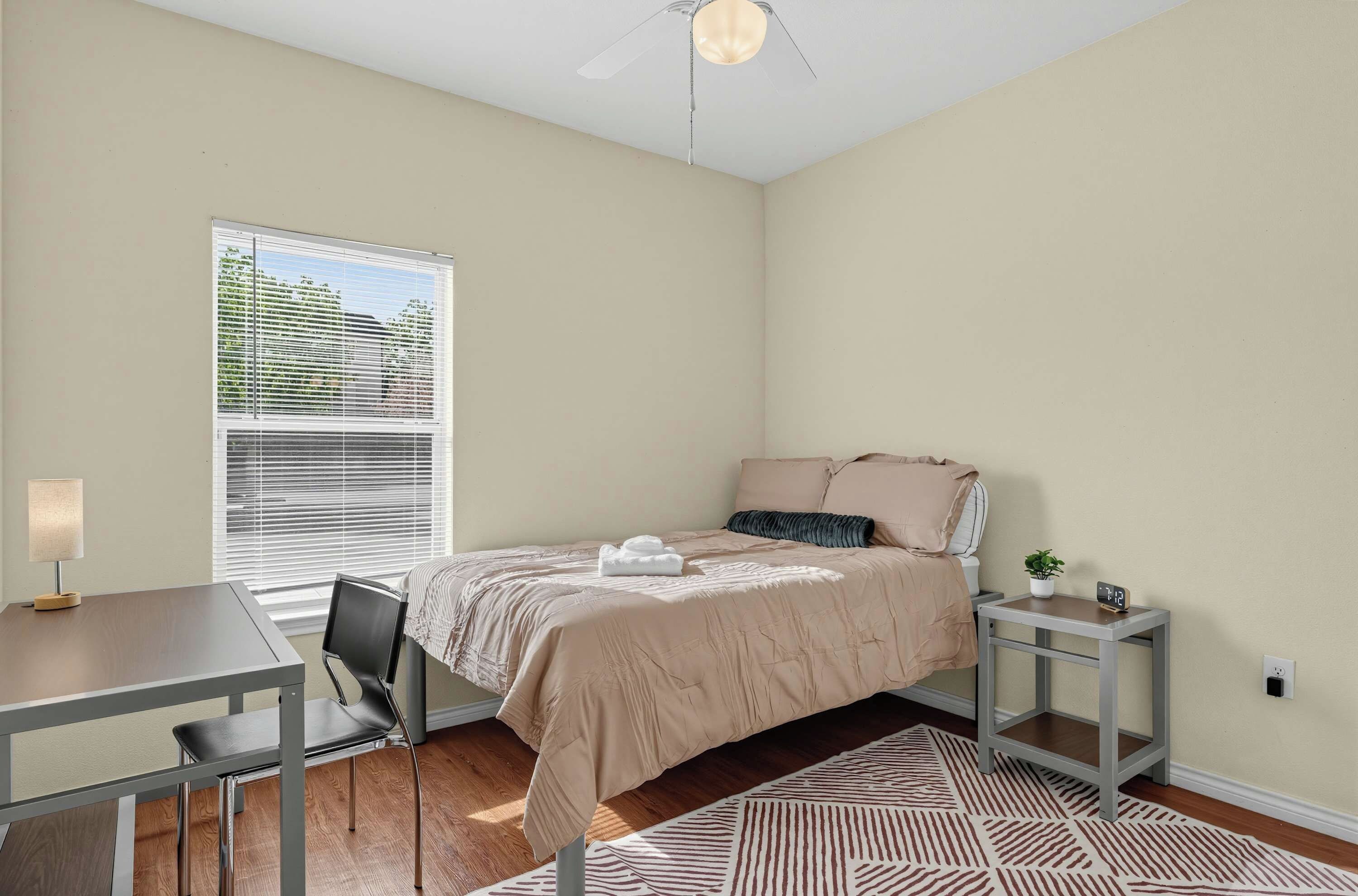 A bedroom with a bed, a desk, a chair, a window with blinds, a ceiling fan, and a patterned rug.