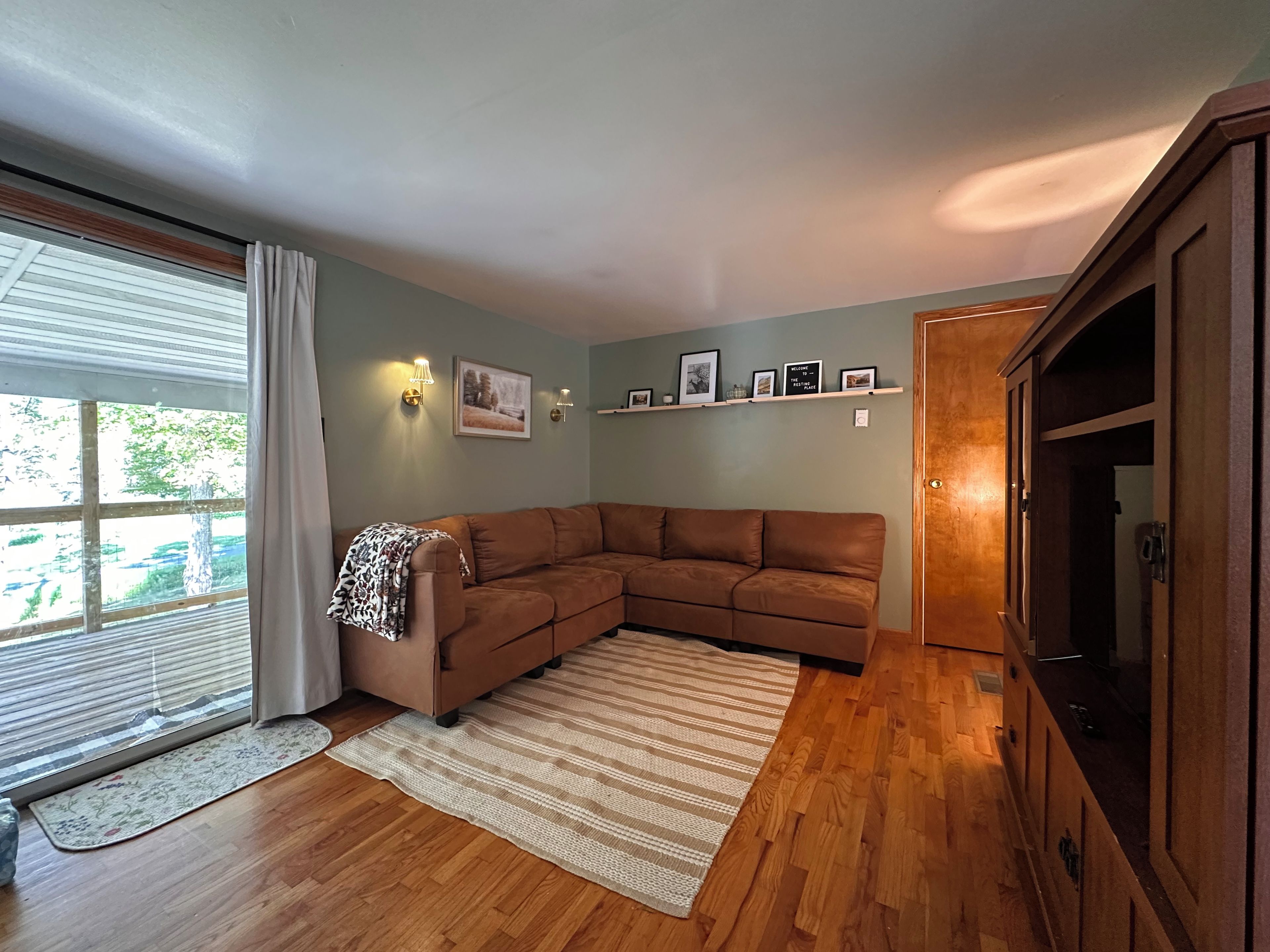 Un salon avec un canapé d’angle marron, un meuble en bois et une fenêtre avec des stores.