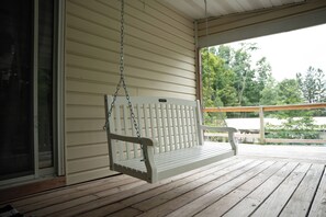 A white porch swing with a 'Kids Only' sign, wooden deck flooring, and a view of greenery through the open porch.