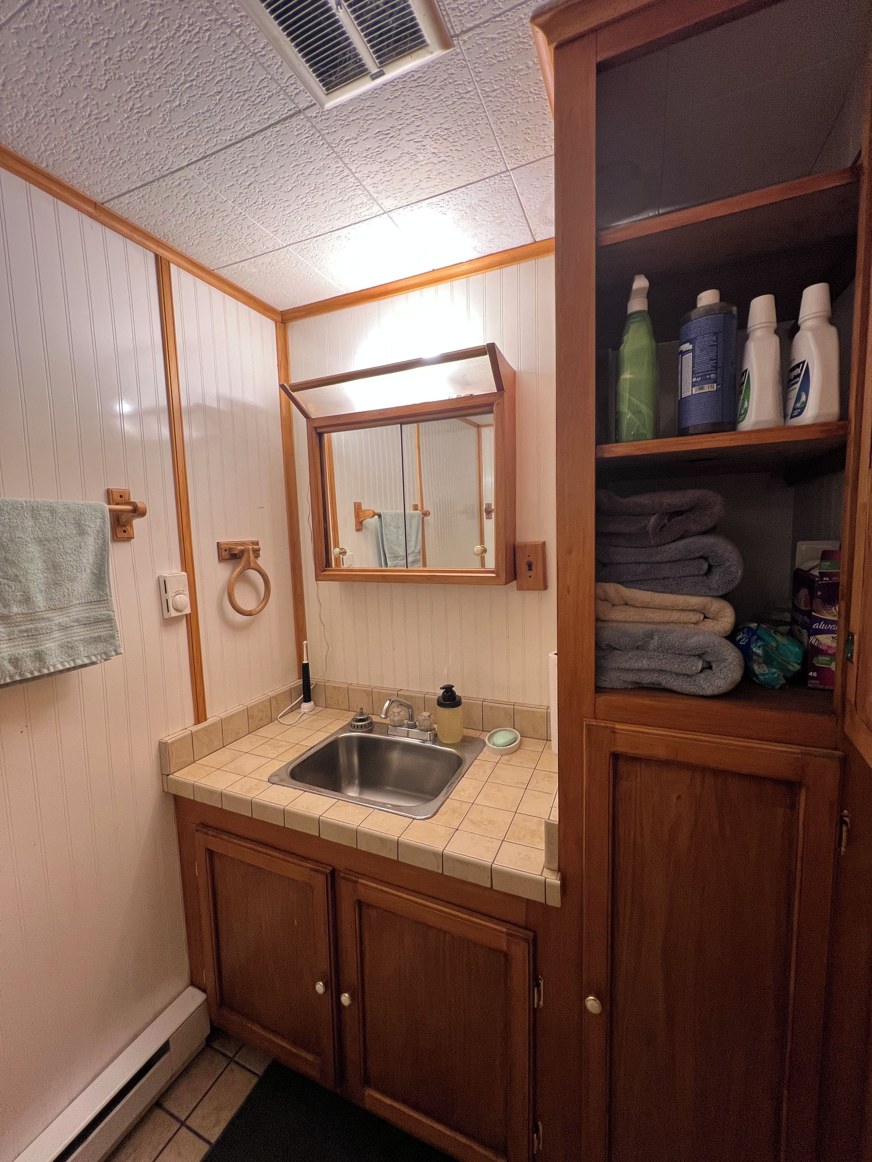 Un baño con lavamanos, espejo, toallero y un mueble con artículos de aseo.