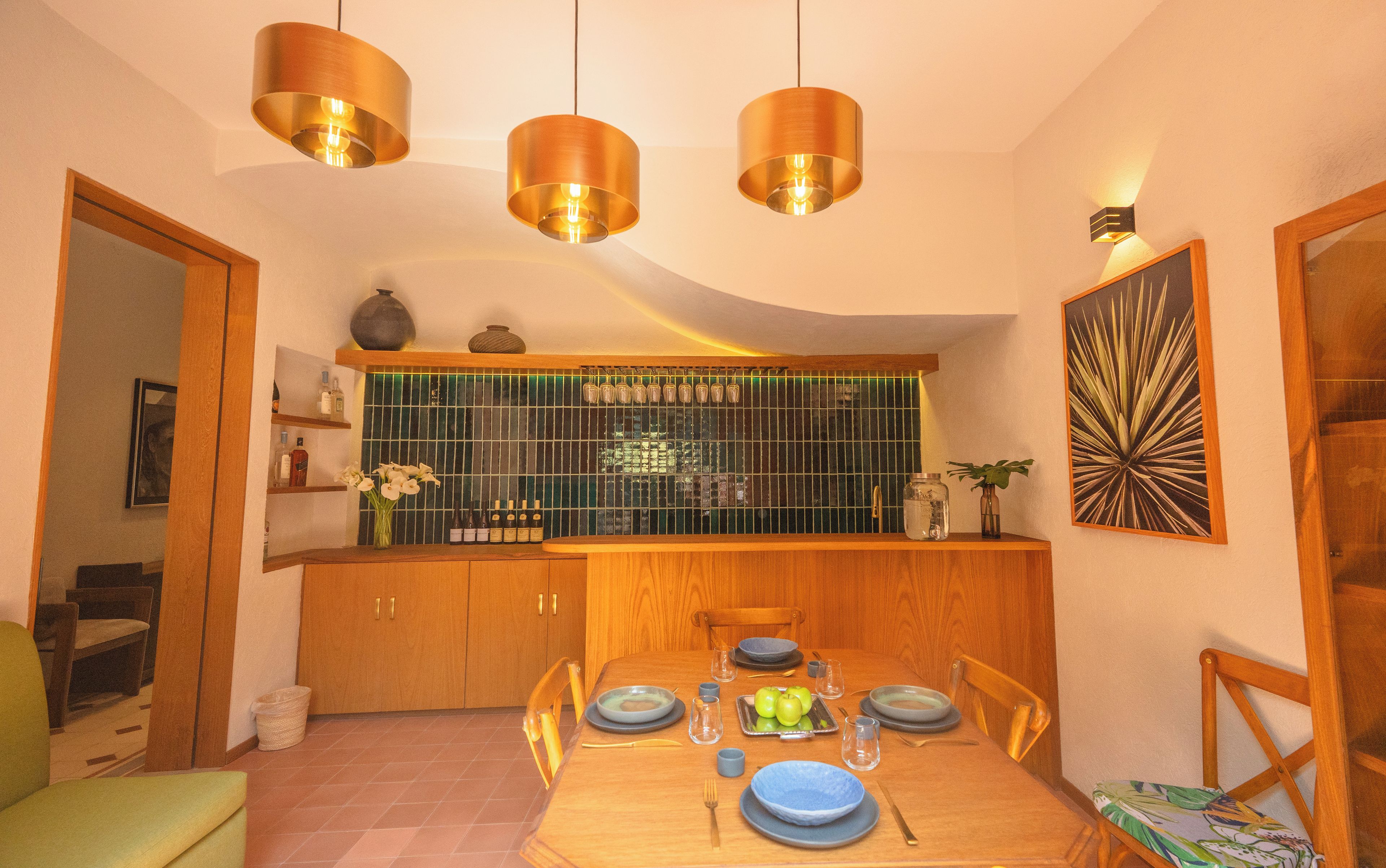 A dining area with a wooden table set for four, a kitchen with a green-tiled backsplash, and pendant lighting.