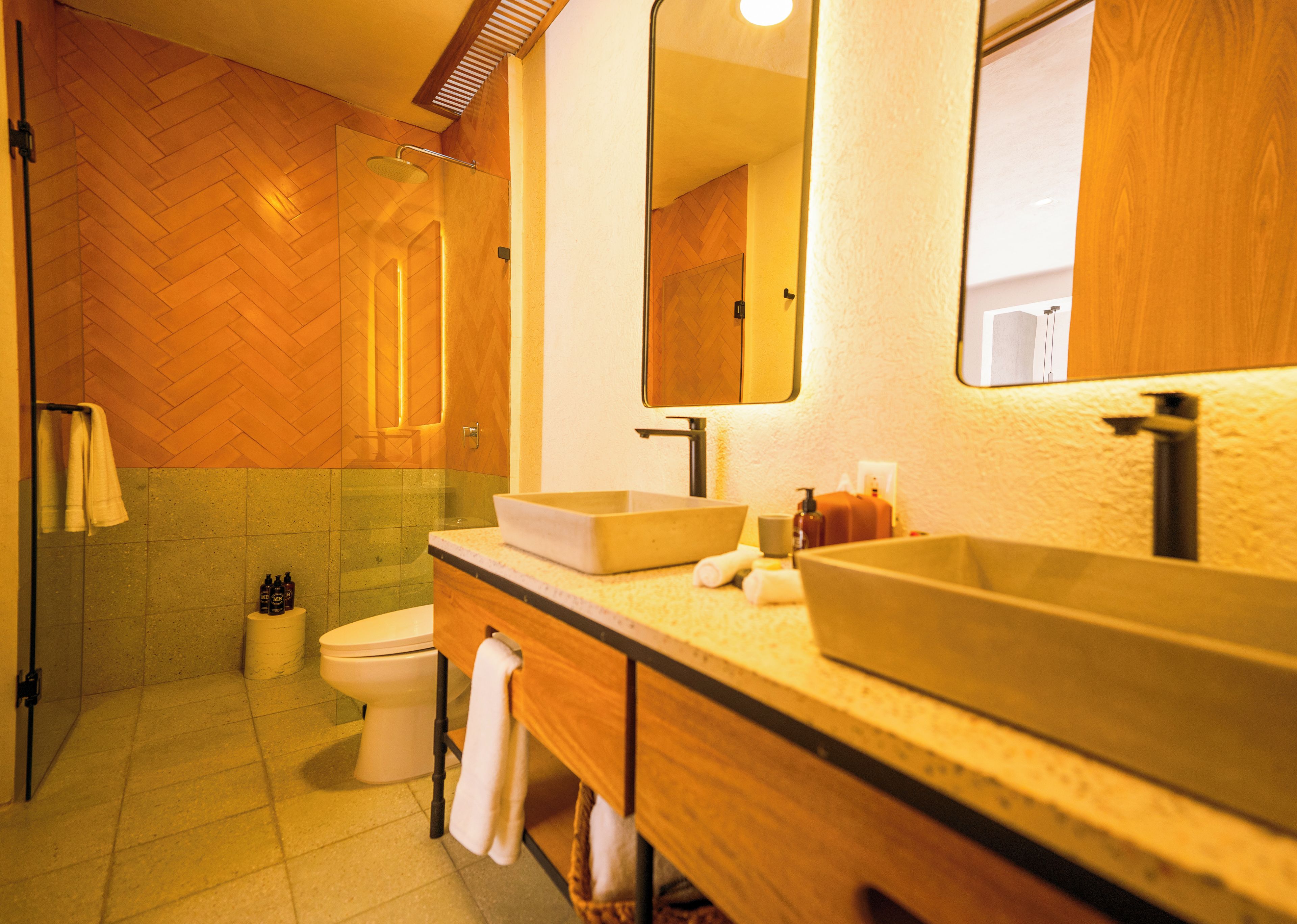 Un baño con piso de madera en patrón de espiga, inodoro blanco, espejo grande y tocador de madera con encimera beige.
