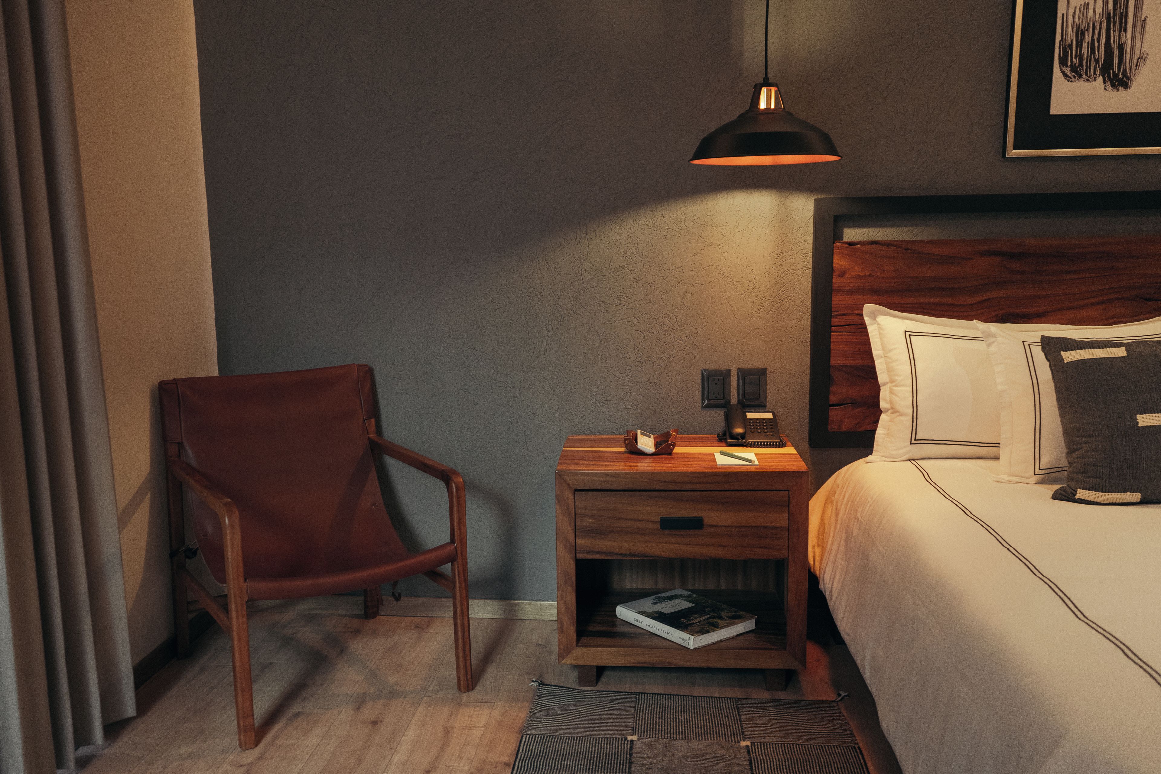 Habitación de hotel con una silla de madera, una mesita de noche con un libro, una lámpara y una cama con almohadas.