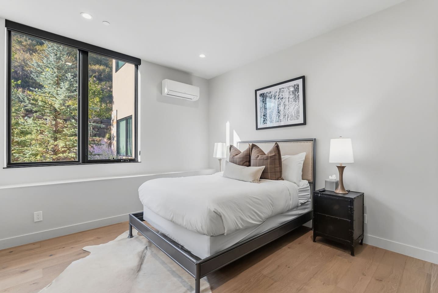 Un dormitorio moderno con cama, mesitas de noche, ventana con vista a árboles y unidad de aire acondicionado.
