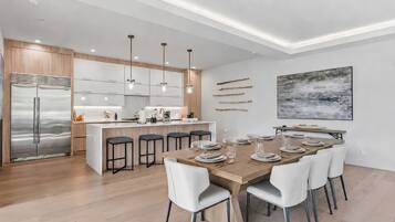 Una cocina moderna con una isla central, electrodomésticos de acero inoxidable, iluminación colgante y un comedor con mesa de madera dispuesta para seis personas.