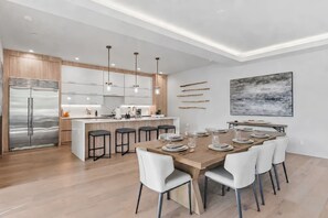 Una cocina moderna con una isla central, electrodomésticos de acero inoxidable, iluminación colgante y un comedor con mesa de madera dispuesta para seis personas.