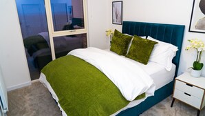 Un dormitorio con cama, ropa de cama verde y blanca, una mesita de noche, una ventana y una puerta.