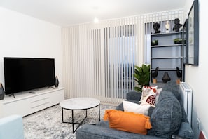 A modern living room with a flat-screen TV, a round coffee table, and a grey sofa with orange cushions.