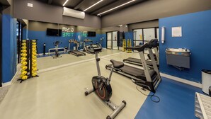 Un gimnasio con diversos equipos de ejercicio, incluyendo cintas de correr y una máquina elíptica, con una pared azul y un reloj digital.