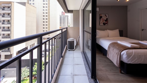 Un balcón con barandilla de metal, una cama con ropa blanca, un cesto de basura y vistas a edificios altos.