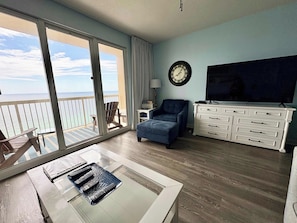 A room with a balcony, a TV, a chair, a table with remotes, and a clock on the wall. - Seychelles Beach Resort 1904 (Panama City Beach)
