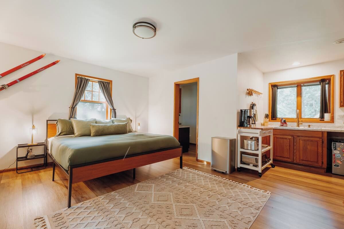 A bedroom with a bed, bedside table, and a nightstand. There is a window with curtains, a door leading to another room, and a kitchen area with cabinets and a countertop.