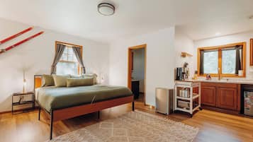 A bedroom with a bed, bedside table, and a nightstand. There is a window with curtains, a door leading to another room, and a kitchen area with cabinets and a countertop.