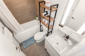 A modern bathroom with a bathtub, toilet, and a shelf unit with toiletries. - Luxury Skyline Condo | Pool • Gym • Steps to SQ1 (Mississauga)