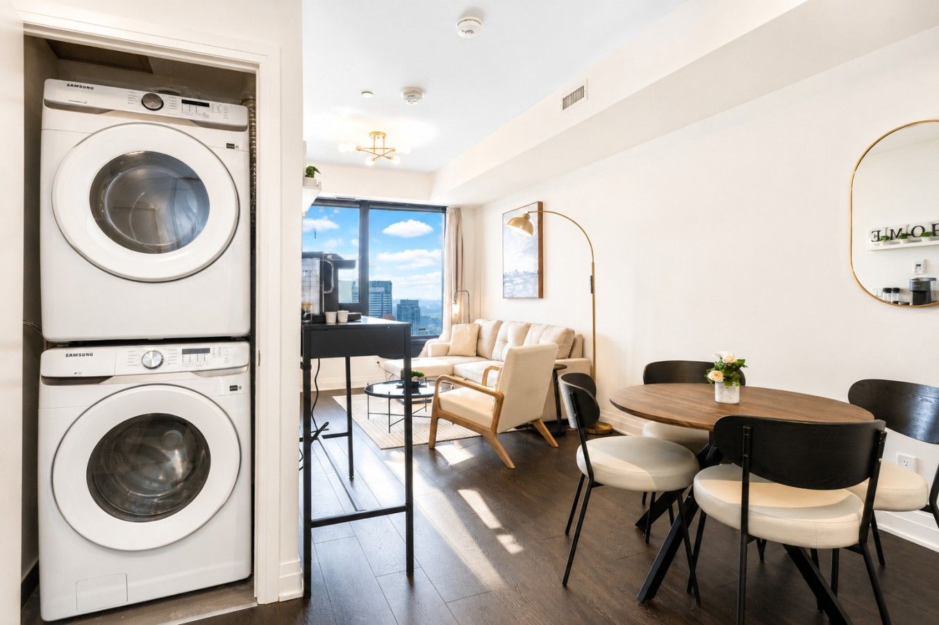 Un appartement moderne équipé d’un lave-linge et d’un sèche-linge, d’un coin repas avec une table ronde et des chaises, d’un canapé et d’une grande fenêtre offrant une vue sur la ville.