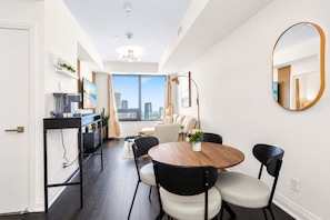A modern living room with a dining area, a large window with a city view, and a round wooden table with four chairs. - Luxury Skyline Condo | Pool • Gym • Steps to SQ1 (Mississauga)