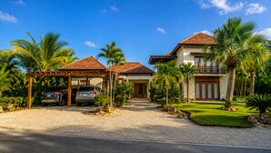 A two-story house with a covered carport, palm trees, and a well-maintained lawn. - Villa CapCana: 5-Bedroom Villa with Pool, Jacuzzi, Beach, and Golf (Punta Cana)