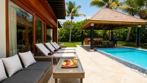 Uma área à beira da piscina com espreguiçadeiras, uma mesa com frutas e bebidas, e um espaço de estar coberto.