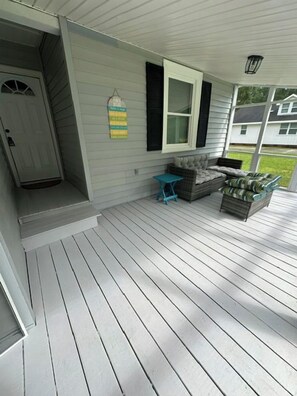 A covered porch with a white railing, a door, and a window with shutters. There is a blue chair and a small table on the porch. - Minutes to Beaches • Self Check-In • Fully Remodeled • Screened Porch • Cozy Fireplace • Washer & Dryer (Wilmington)