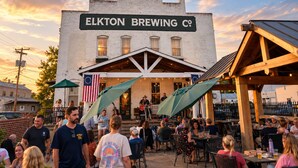 Ein Außenbereich mit Tischen und Stühlen vor einem Gebäude, an dem ein Schild mit der Aufschrift 'ELKTON BREWING CO' zu sehen ist.