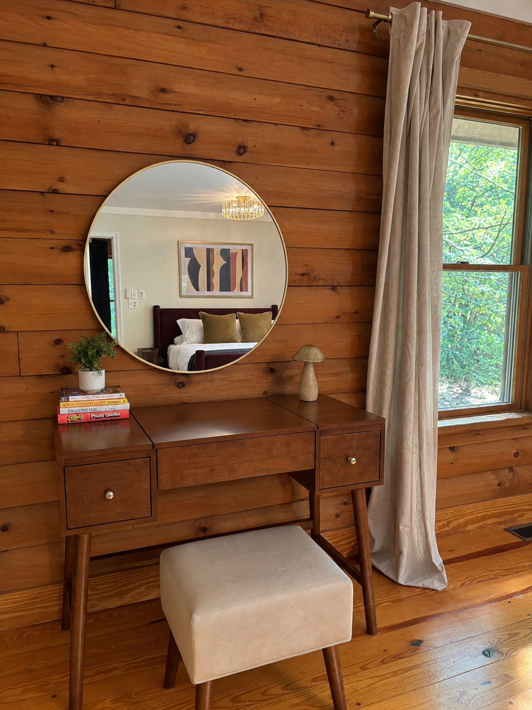 A wooden-paneled room with a wooden vanity, a round mirror, a small stool, and a window with a curtain.
