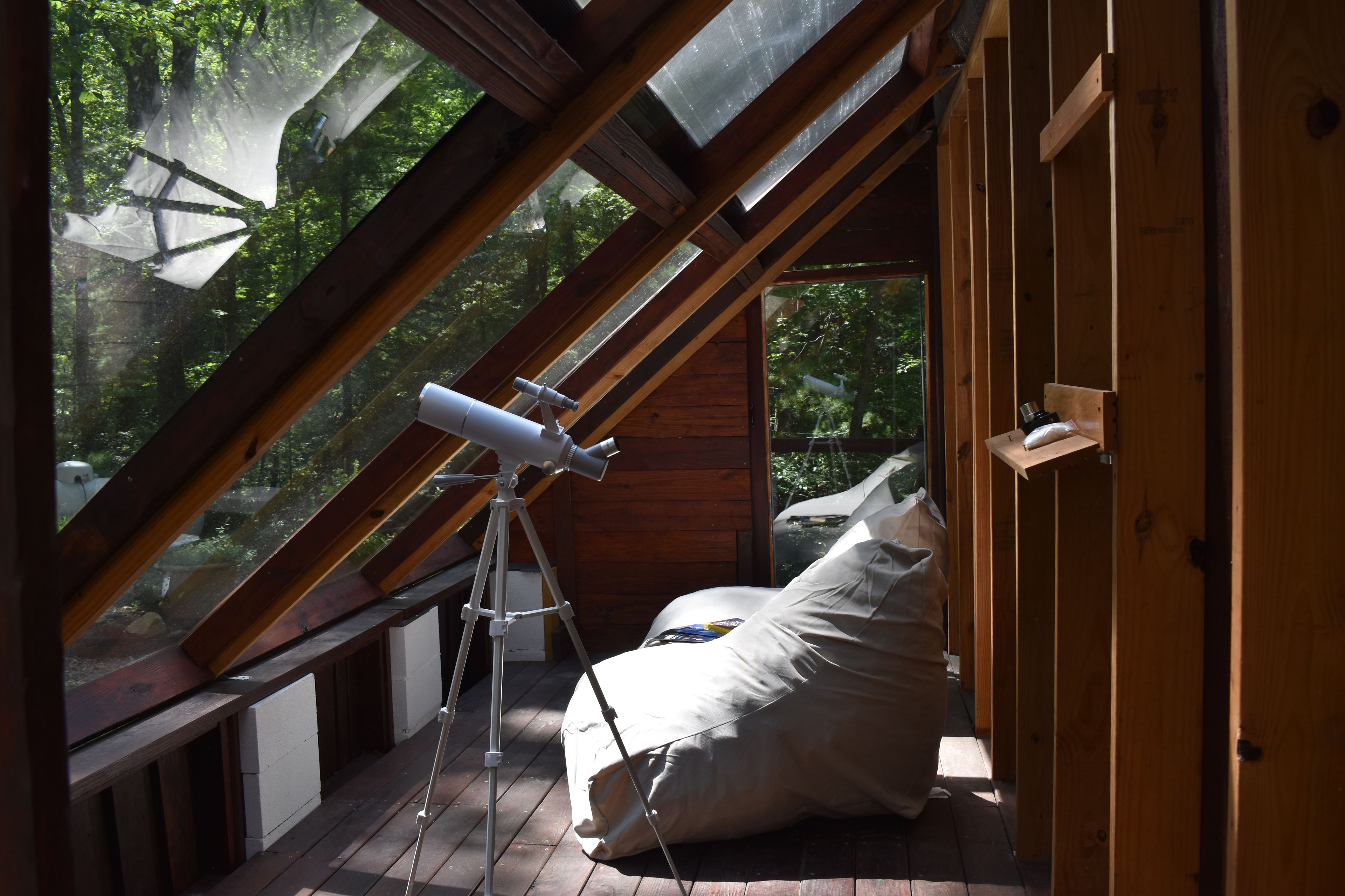 A telescope set up in a wooden attic room with a view of a forest through a skylight.