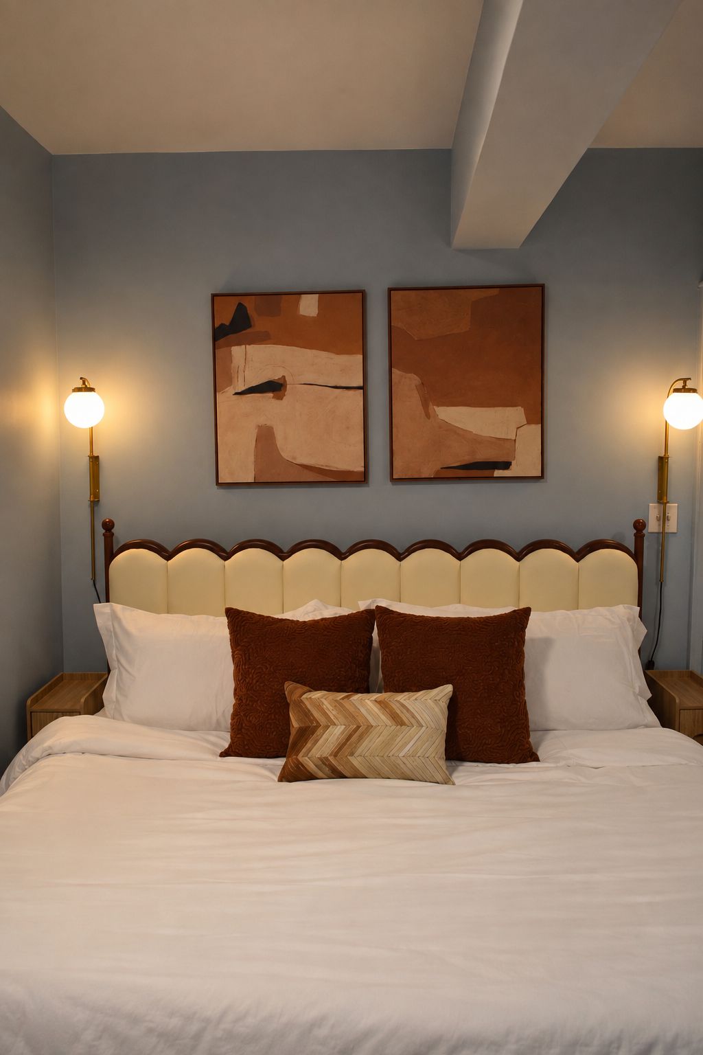 Una cama con sábanas blancas y almohadas marrones, flanqueada por dos cuadros colgados en la pared.