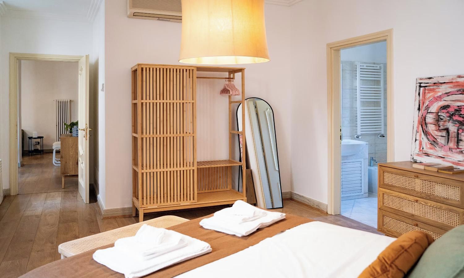 A hotel room with a bed, a wooden wardrobe, a mirror, and a painting on the wall.