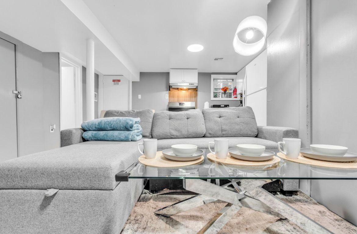 A modern living room with a glass coffee table, grey sofa, and a kitchen area in the background.