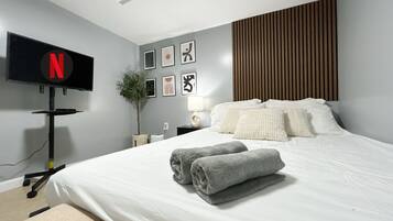 A modern bedroom with a large bed, a flat-screen TV mounted on the wall, a bedside table with a lamp, and a potted plant.
