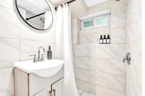 Kylpyhuone, jossa on valkoinen pesuallas, peili, suihkuverho ja hylly, jossa on hygieniatuotteita.