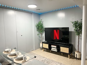 Huoneessa on televisio, jossa näkyy Netflixin logo, ruokapöytä, jossa on lautasia ja kuppeja, sekä kaksi ruukkukasvia.