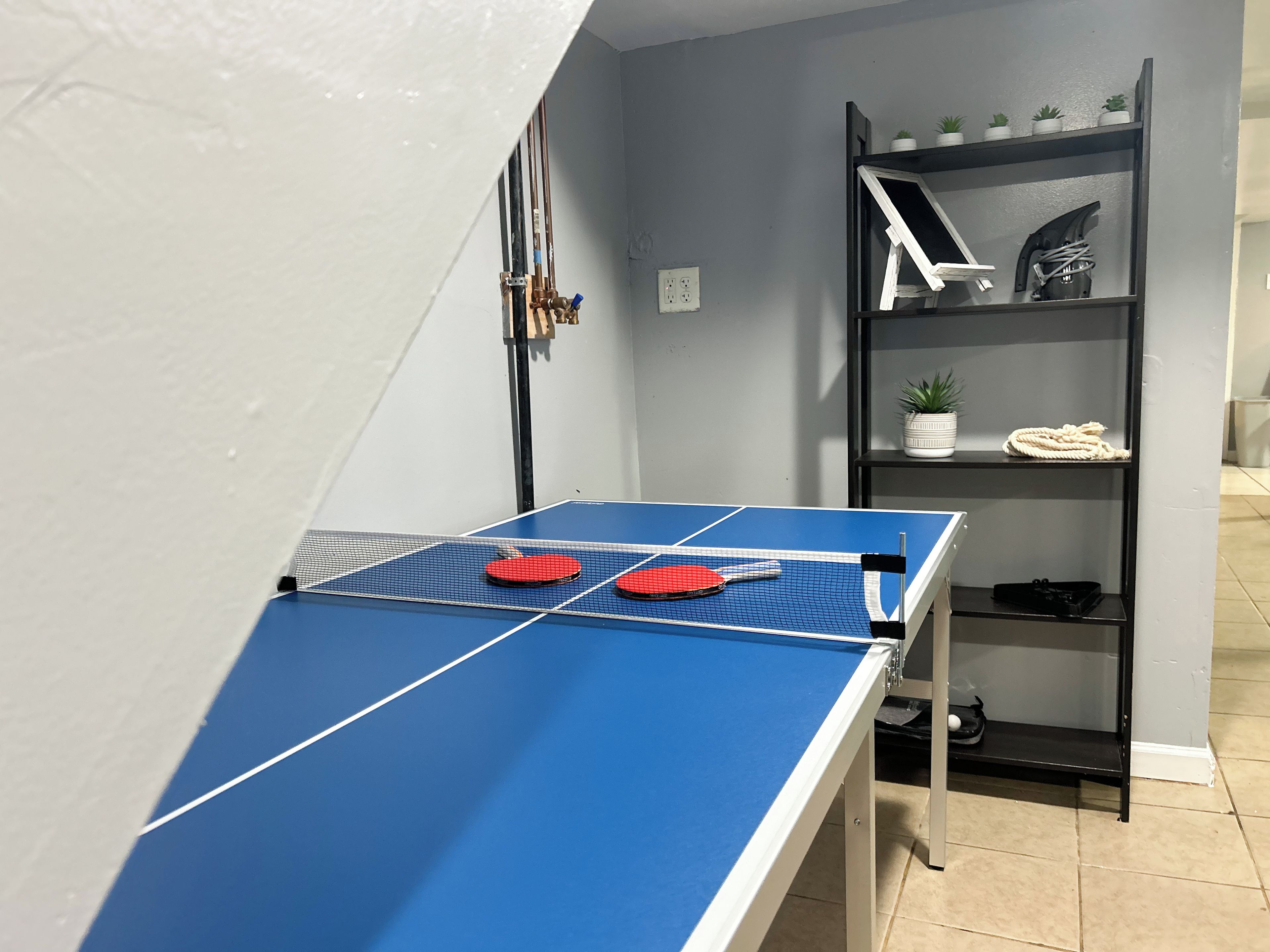 Une table de ping-pong avec deux raquettes rouges, une étagère en métal noir ornée de plantes et une prise de courant blanche.