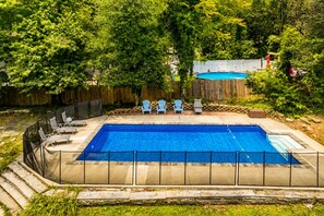 A rectangular swimming pool with a stone border and a wooden deck surrounding it. There are lounge chairs and a small table near the pool.