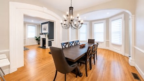 A dining area with a wooden table and chairs, a chandelier, and large windows with blinds.