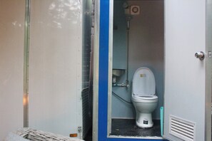 Siirrettävä wc, jossa on kansi, suihkutila ja metallinen ovenkarmi.