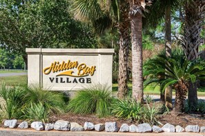 A sign for Hidden Bay Village with a landscaped area featuring palm trees and shrubs. - Townhome with Shared Pool, Wifi & Full Kitchen (Navarre)