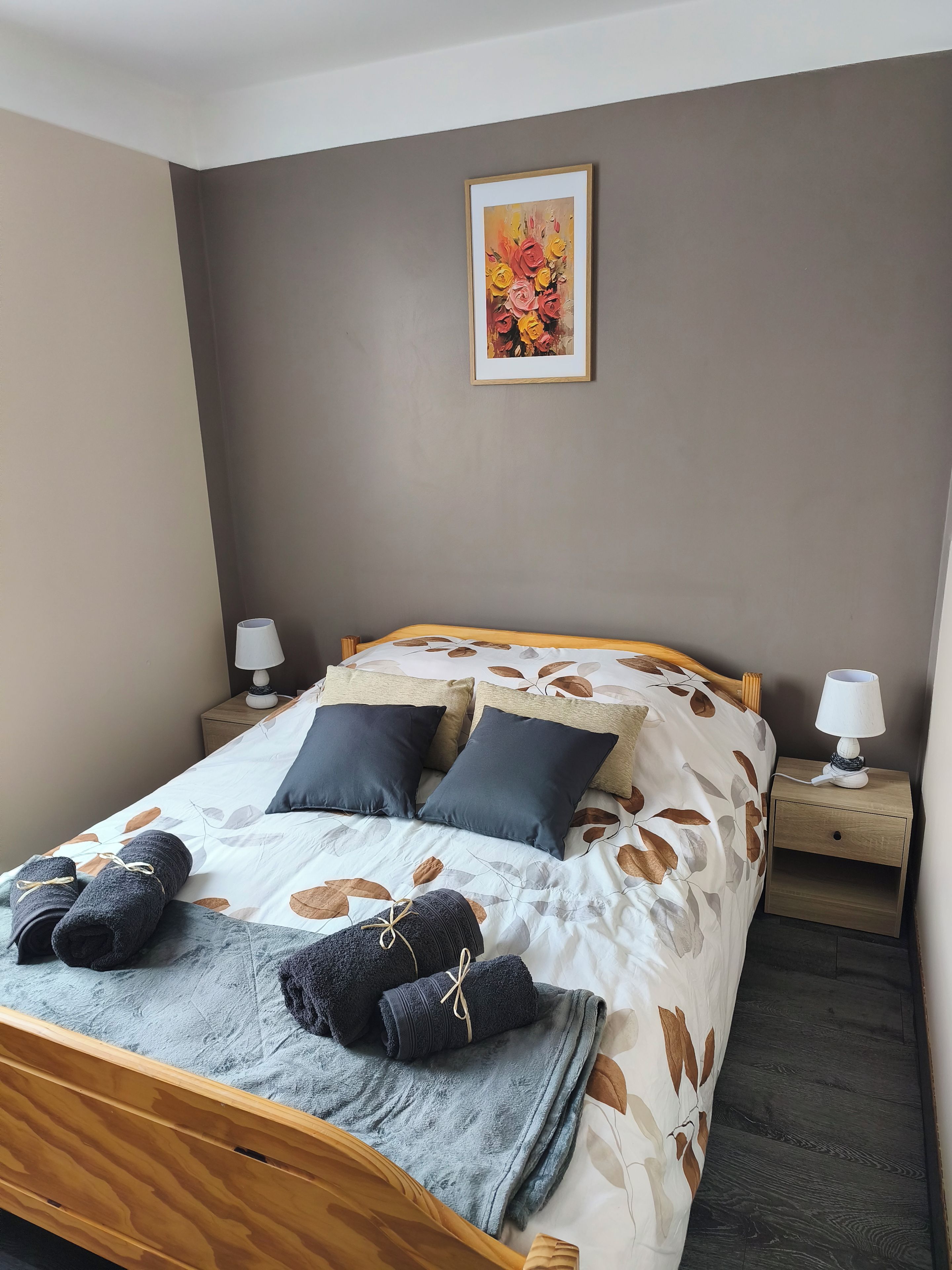 A neatly made bed with a floral-patterned duvet, two pillows, and rolled towels. There is a framed artwork on the wall above the bed.