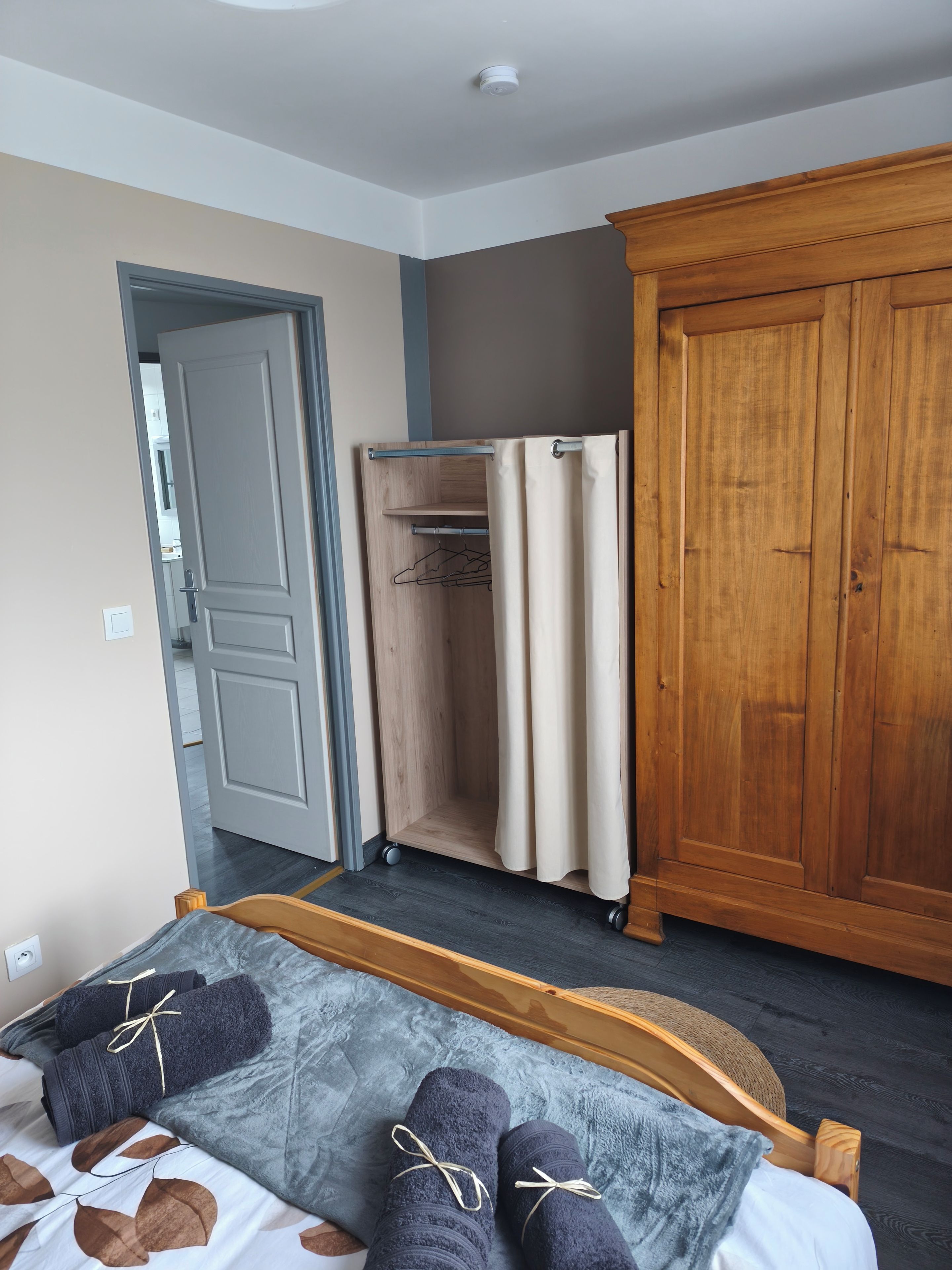 Une chambre à coucher comprenant un lit, une armoire en bois et une autre armoire fermée par un rideau, ainsi qu’une porte donnant sur une autre pièce.