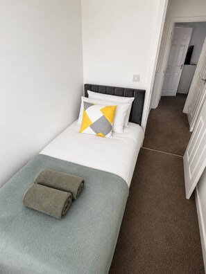 A single bed with a grey headboard, white bedding, and a patterned throw blanket.