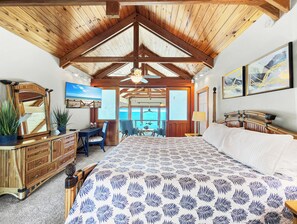 A bedroom with a large bed, wooden ceiling beams, a desk with a chair, a dresser, and a view of the ocean through a door. - Island Dream  Beachfront Plantation Home  2 Kings + Bunk Room  Sleeps 10 (Eastpoint)