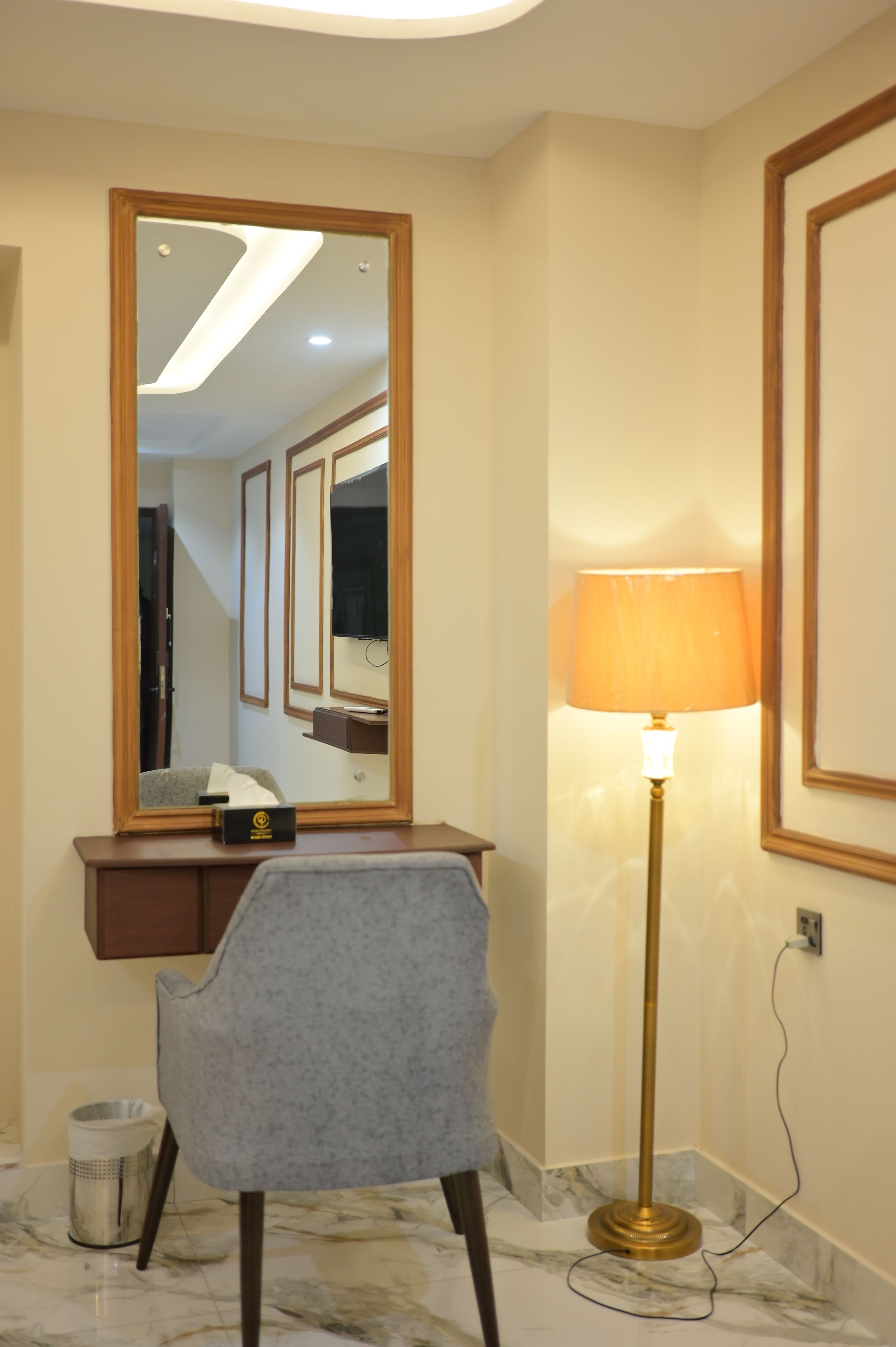 Habitación de hotel con piso de mármol, un escritorio con silla, una lámpara de pie y un espejo grande.